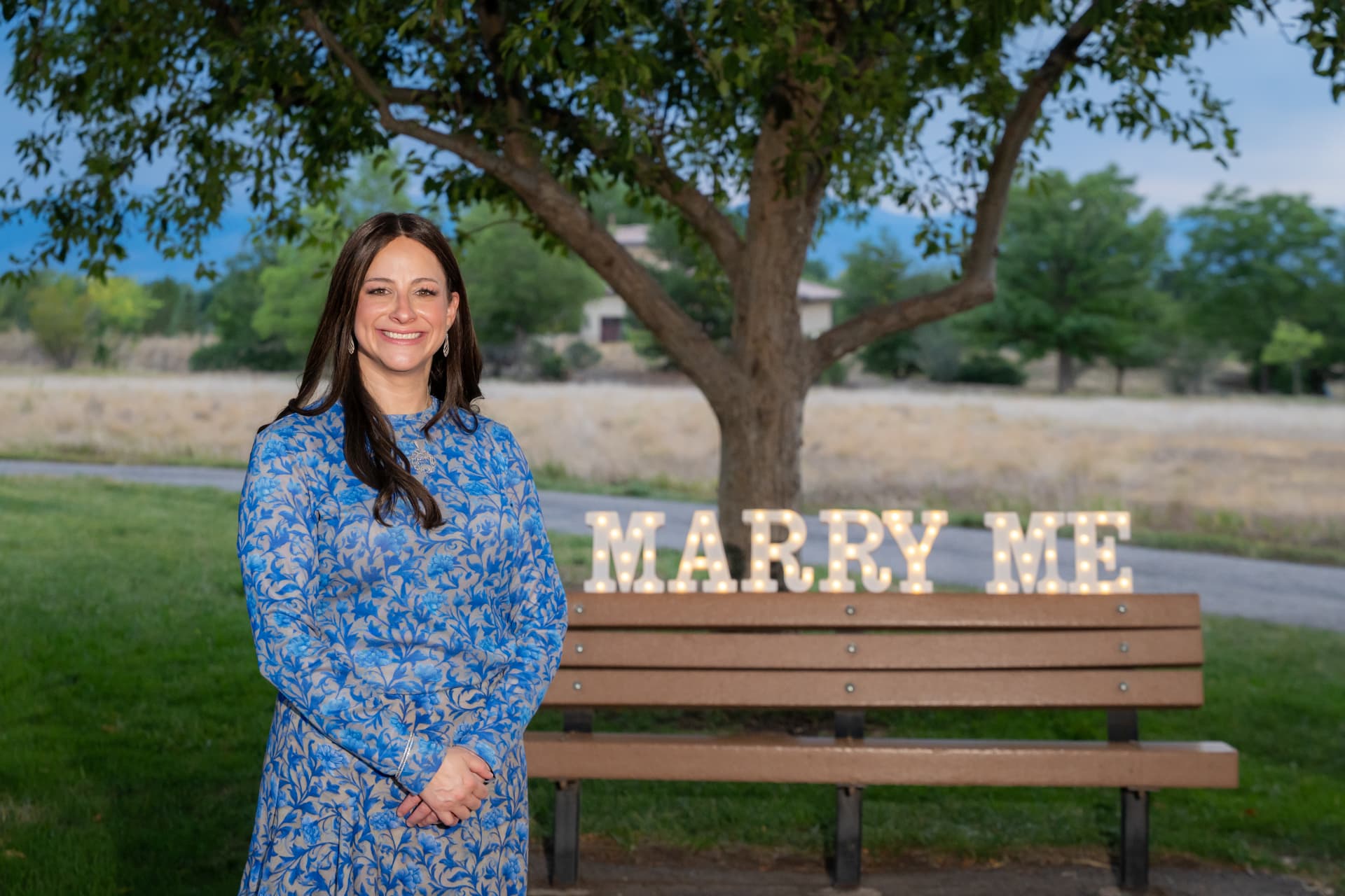A couple celebrates a marriage proposal at dusk in a park, with glowing 'MARRY ME' marquee letters on a wooden bench behind the woman, symbolizing the joy and intention behind premarital counseling.